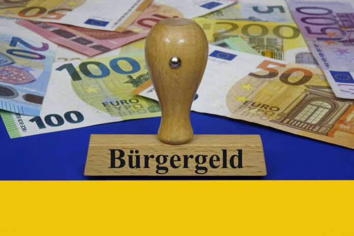 Ein Stempel mit dem Aufdruck "Bürgergeld" liegt auf einer ukrainischen Flagge. Hier liegt auch eine Menge Bargeld, das den staatlichen Zuschuss für Flüchtlinge symbolisiert. Es sind 5-Euro, 10-Euro, 20-Euro, 50-Euro-, 100-Euro, 200-Euro und ein 500-Euro-Schein.