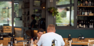 Ein Mann und eine Frau sitzen an einem Tisch in einem Restaurant und essen ihr bestelltes Essen. Sie unterhalten sich. Man sieht weitere leere Stühle und Tische sowie im Hintergrund an einem Tresen eine junge Frau.