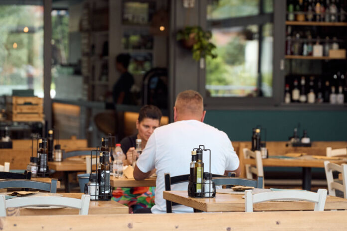 Ein Mann und eine Frau sitzen an einem Tisch in einem Restaurant und essen ihr bestelltes Essen. Sie unterhalten sich. Man sieht weitere leere Stühle und Tische sowie im Hintergrund an einem Tresen eine junge Frau.