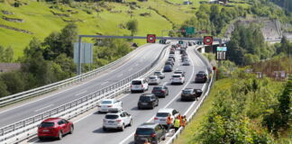 Vor einem Tunnel hat sich in einer Richtung auf der Autobahn ein Stau gebildet. Die Autobahn ist umgeben von Bergen. Es scheint Sommer zu sein und die Sonne scheint.