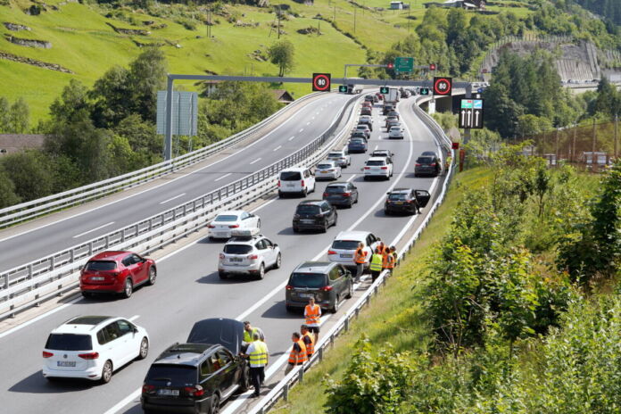 Vor einem Tunnel hat sich in einer Richtung auf der Autobahn ein Stau gebildet. Die Autobahn ist umgeben von Bergen. Es scheint Sommer zu sein und die Sonne scheint.