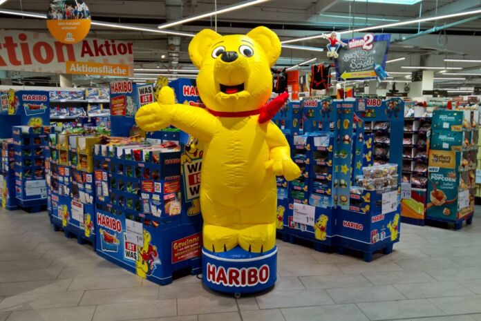 Ein großer aufblasbarer Haribo-Bär steht neben einem großen Regal mit vielen Süßigkeiten des Herstellers Haribo. Mehrere Regale stehen hintereinander und nebeneinander in einem Supermarkt oder Discounter.