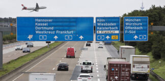 Eine mehrspurige deutsche Autobahn mit regem Verkehr. Dicht über der Fahrbahn fliegt ein großes Passagier-Flugzeug. Es landet auf dem Flughafen in unmittelbarer Nähe.