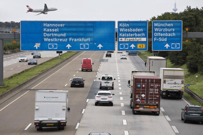 Eine mehrspurige deutsche Autobahn mit regem Verkehr. Dicht über der Fahrbahn fliegt ein großes Passagier-Flugzeug. Es landet auf dem Flughafen in unmittelbarer Nähe.