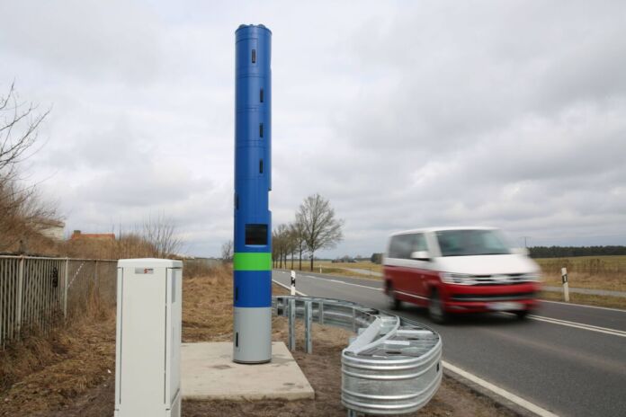 Eine große blaue Säule steht am Rand einer Landstraße, auf der gerade ein VW-Bus fährt. Die blaue Säule erfasst jedes Auto, das an ihr vorbeifährt.