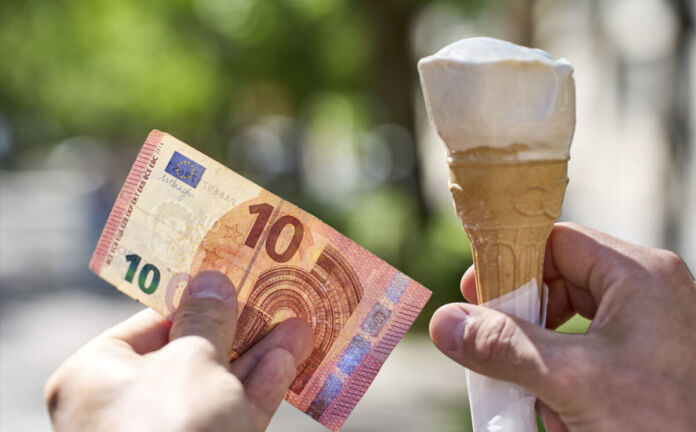 Es ist Sommer und eine Hand hält eine Kugel Eis in der Eiswaffel. Die andere Hand hält einen Zehn-Euro-Schein. Im Hintergrund kann man Bäume sowie Gebäude erkennen.