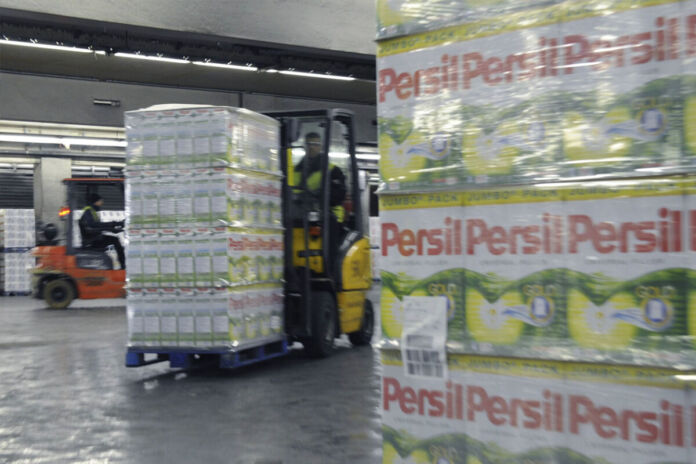 Ein Blick in eine Fabrik, die Waschmittel herstellt. Es handelt sich um die bekannte Marke Persil. Auf mehreren Gabelstaplern befindet sich das abgepackte Waschpulver auf Paletten übereinander gestapelt und wird umhergefahren.