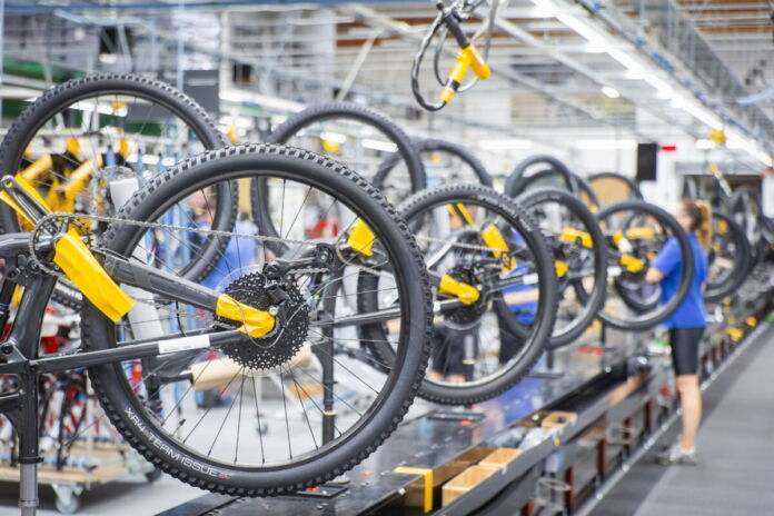 In einer hellen Werkstatt eines Fahrradherstellers werden am Fließband dutzende Räder hergestellt. Die schwarzen Reifen sieht man in einer Nahaufnahme, während auch an der Decke die Räder des Herstellers hängen.