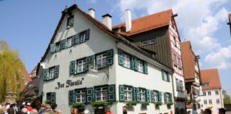 Vor einer großen bunten Menschenmenge erhebt sich ein wunderschönes Altbau-Gasthaus mit rotem Dach, Giebel, grünen Fensterläden und einer weißen Fassade sowie schönem Dekor.