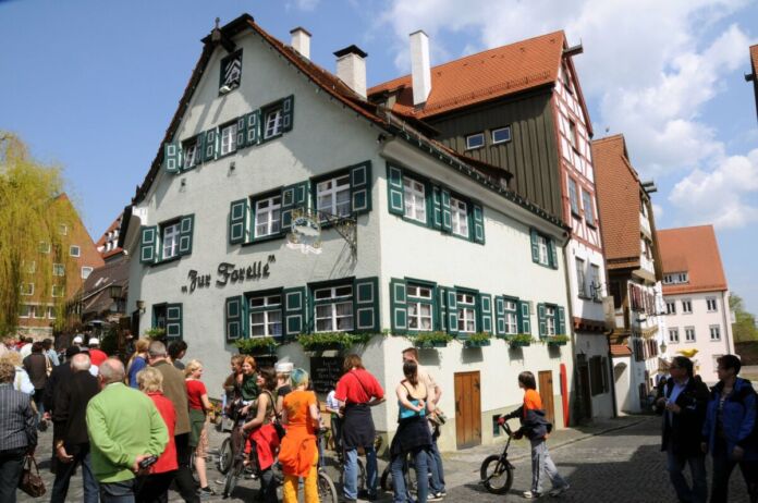 Vor einer großen bunten Menschenmenge erhebt sich ein wunderschönes Altbau-Gasthaus mit rotem Dach, Giebel, grünen Fensterläden und einer weißen Fassade sowie schönem Dekor.