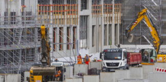 Auf einer großen Baustelle herrscht großer Trubel. Verschiedene Fahrzeuge parken vor dem Rohbau, Arbeiter mit Warnwesten koordinieren die Bauarbeiten. Man sieht im Vordergrund große Betonblöcke als Stützpfeiler für das Fundament