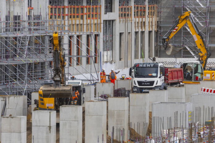 Auf einer großen Baustelle herrscht großer Trubel. Verschiedene Fahrzeuge parken vor dem Rohbau, Arbeiter mit Warnwesten koordinieren die Bauarbeiten. Man sieht im Vordergrund große Betonblöcke als Stützpfeiler für das Fundament