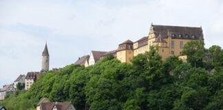 Auf einem bewaldeten Hügel in einer kleinen Stadt steht eine große, gut erhaltene Residenz-Burg. Die gelben Fassaden und roten Ziegeldächer strahlen in der Sonne und ganz links steht einsam ein kleines Türmchen.