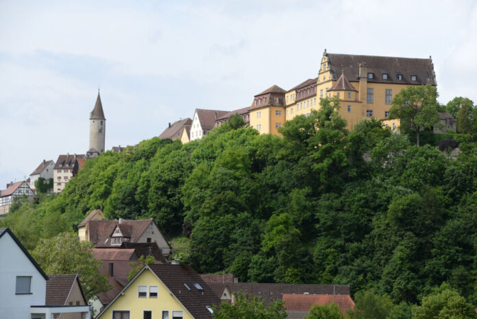 Auf einem bewaldeten Hügel in einer kleinen Stadt steht eine große, gut erhaltene Residenz-Burg. Die gelben Fassaden und roten Ziegeldächer strahlen in der Sonne und ganz links steht einsam ein kleines Türmchen.