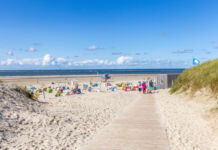 An einem heiteren Tag befinden sich viele Menschen mit Sonnenliegen und Sonnenschirmen am Strand. Dieser ist groß und voller Sand, einladend und zieht sich lang am Meer entlang.