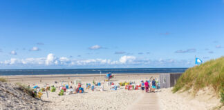 An einem heiteren Tag befinden sich viele Menschen mit Sonnenliegen und Sonnenschirmen am Strand. Dieser ist groß und voller Sand, einladend und zieht sich lang am Meer entlang.