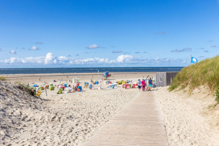 An einem heiteren Tag befinden sich viele Menschen mit Sonnenliegen und Sonnenschirmen am Strand. Dieser ist groß und voller Sand, einladend und zieht sich lang am Meer entlang.