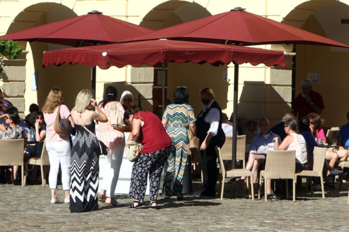 Vor einem Restaurant mit roten Sonnenschirmen an einem schönen Tag im Sommer mitten in der Stadt warten mehrere Menschen in einer Schlange darauf, bedient und an den Tisch gesetzt zu werden.