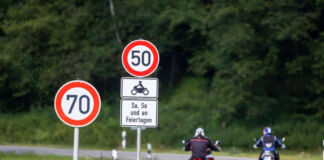 Ein Blick auf eine Landstraße, welche eine Kurve schlägt. Zwei Motorradfahrer fahren die Straße entlang. Im Hintergrund erkennt man eine grüne Wiese und einen grünen Wald. Im Vordergrund stehen zwei Verkehrsschilder mit einem Tempolimit von 70 und einem Tempolimit von 50 an Wochenenden und Feiertagen für Biker.