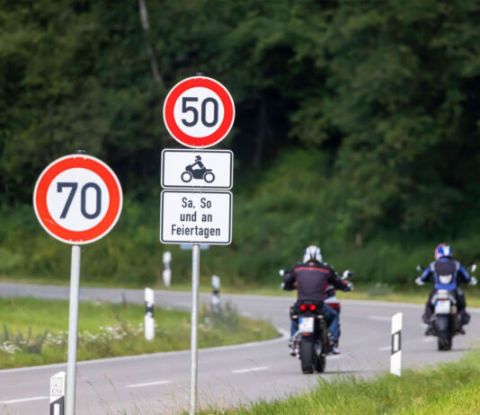 Ein Blick auf eine Landstraße, welche eine Kurve schlägt. Zwei Motorradfahrer fahren die Straße entlang. Im Hintergrund erkennt man eine grüne Wiese und einen grünen Wald. Im Vordergrund stehen zwei Verkehrsschilder mit einem Tempolimit von 70 und einem Tempolimit von 50 an Wochenenden und Feiertagen für Biker.