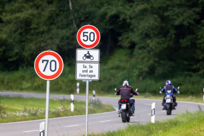 Ein Blick auf eine Landstraße, welche eine Kurve schlägt. Zwei Motorradfahrer fahren die Straße entlang. Im Hintergrund erkennt man eine grüne Wiese und einen grünen Wald. Im Vordergrund stehen zwei Verkehrsschilder mit einem Tempolimit von 70 und einem Tempolimit von 50 an Wochenenden und Feiertagen für Biker.