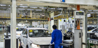 Ein Mitarbeiter steht in einer Fertigungshalle in einem Werk von VW und schließt gerade die Motorhaube. Er trägt einen Blaumann und weiße Handschuhe.