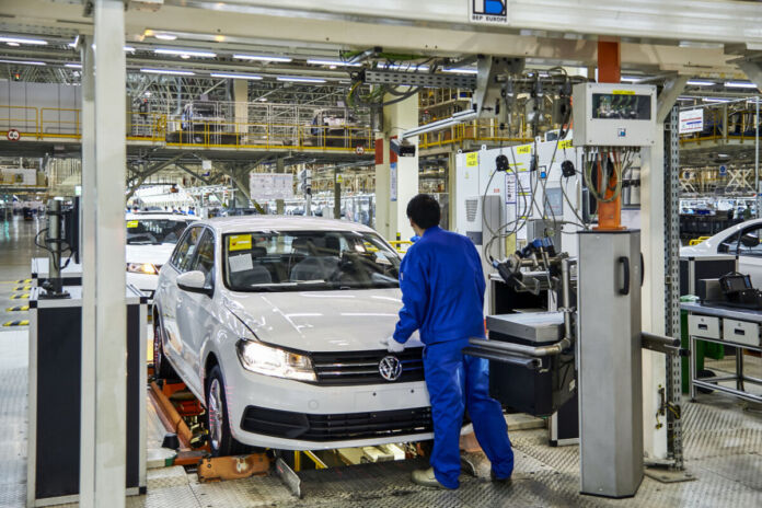 Ein Mitarbeiter steht in einer Fertigungshalle in einem Werk von VW und schließt gerade die Motorhaube. Er trägt einen Blaumann und weiße Handschuhe.