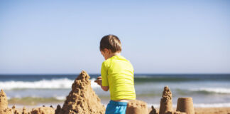 Ein Kind baut eine Sandburg an einem Strand. Im Hintergrund befindet sich das Meer und die Wellen. Das spielende Kind trägt ein gelbes T-Shirt und eine blaue Badehose.