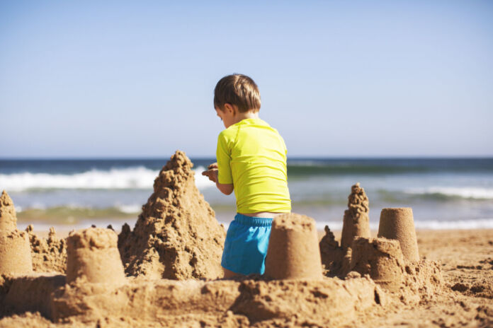 Ein Kind baut eine Sandburg an einem Strand. Im Hintergrund befindet sich das Meer und die Wellen. Das spielende Kind trägt ein gelbes T-Shirt und eine blaue Badehose.