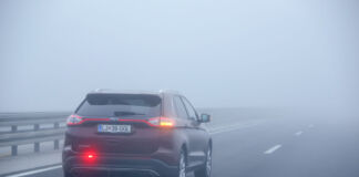 Ein Auto fährt auf einer zweispurigen Straße auf der Überholspur, der linken Seite. Es ist sehr nebelig und die Sicht eingeschränkt. Die Nebelschlussleuchte und der rechte Blinker sind auf der rückwärtigen Seite des Fahrzeugs eingeschaltet.