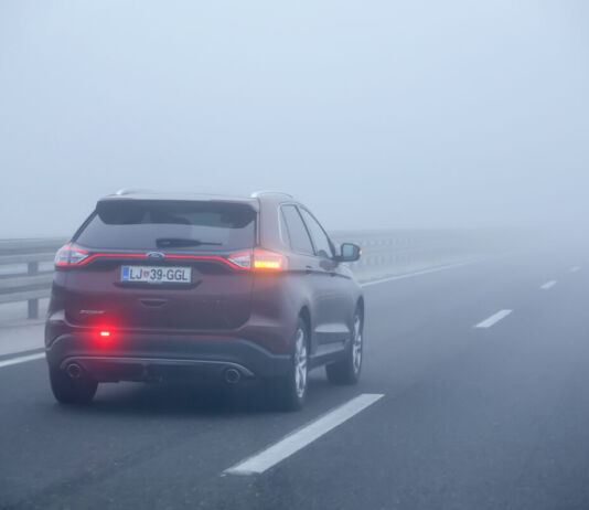 Ein Auto fährt auf einer zweispurigen Straße auf der Überholspur, der linken Seite. Es ist sehr nebelig und die Sicht eingeschränkt. Die Nebelschlussleuchte und der rechte Blinker sind auf der rückwärtigen Seite des Fahrzeugs eingeschaltet.