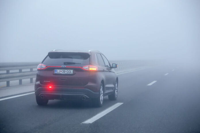 Ein Auto fährt auf einer zweispurigen Straße auf der Überholspur, der linken Seite. Es ist sehr nebelig und die Sicht eingeschränkt. Die Nebelschlussleuchte und der rechte Blinker sind auf der rückwärtigen Seite des Fahrzeugs eingeschaltet.