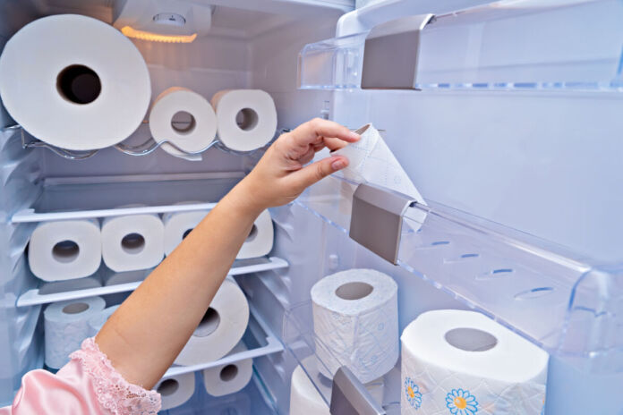 Die Hand einer Frau nimmt gerade eine angefangene Toilettenpapier-Rolle aus der Kühlschranktür. In jedem Fach im Kühlschrank befinden sich viele Rollen Toilettenpapier.