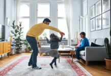 Ein Vater tanzt und tobt mit seiner Tochter in der Wohnung. Die Mutter sitzt auf dem Sofa und schaut den beiden zu.