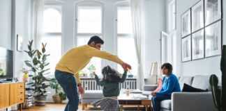 Ein Vater tanzt und tobt mit seiner Tochter in der Wohnung. Die Mutter sitzt auf dem Sofa und schaut den beiden zu.