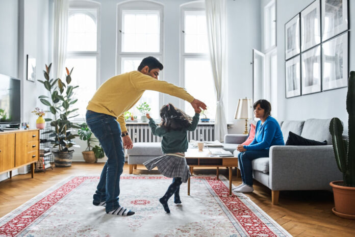 Ein Vater tanzt und tobt mit seiner Tochter in der Wohnung. Die Mutter sitzt auf dem Sofa und schaut den beiden zu.