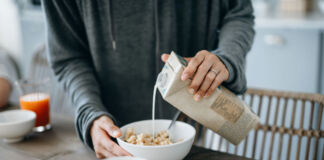 Eine Frau im Pulli hält eine Packung vegane Milch in der Hand und gibt sie zu einer Schüssel mit Müsli. Die Frau bereitet ihr Frühstück mit Milch zu.