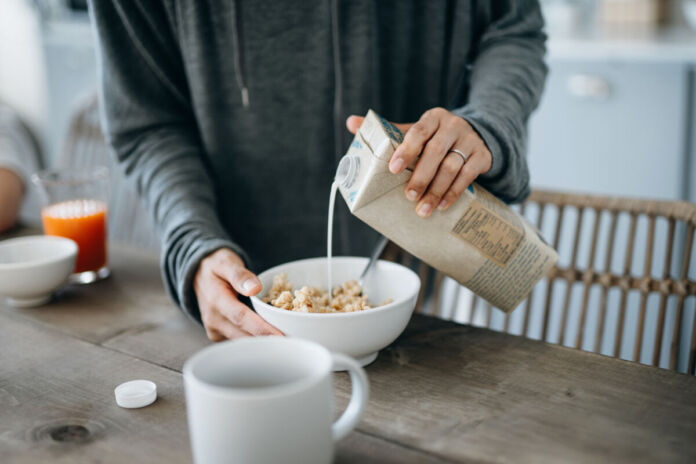 Eine Frau im Pulli hält eine Packung vegane Milch in der Hand und gibt sie zu einer Schüssel mit Müsli. Die Frau bereitet ihr Frühstück mit Milch zu.