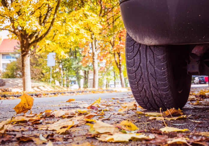 Die Nahaufnahme eines Autoreifens, der auf herbstlichen Straßen fährt. Orange Blätter liegen auf dem Boden. Es herrscht Rutschgefahr im Herbst. Im Hintergrund stehen gelbe Bäume.