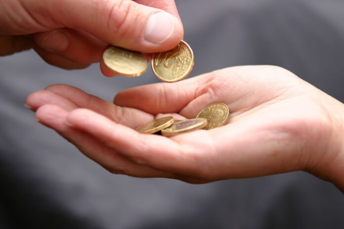 Eine Person hält ihre Hand offen, in der sich schon eine 1-Euro-Münze, ein 50-Cent-Stück und eine 20-Cent-Münze befinden. Die Hand einer anderen Person hält zwei 20-Cent-Münzen und ist gerade im Begriff, eine davon in die Hand der anderen Person zu legen.