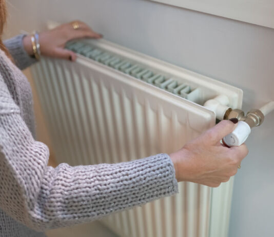 Eine Frau befindet sich offenbar in ihrer Wohnung und dreht die Heizung mit der rechten Hand auf. Mit der linken fühlt sie die Temperatur.