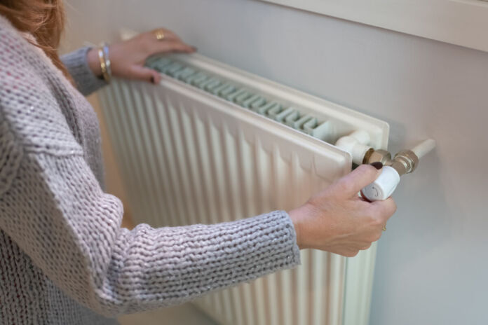 Eine Frau befindet sich offenbar in ihrer Wohnung und dreht die Heizung mit der rechten Hand auf. Mit der linken fühlt sie die Temperatur.