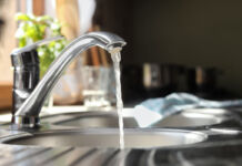 Ein Blick in eine Küche, direkt auf eine Spüle. Der Wasserhahn ist angeschaltet und es fließt klares Leitungswasser in das Waschbecken. Im Hintergrund kann man ein Geschirrhandtuch und einen Topf mit Küchenkräutern erkennen.