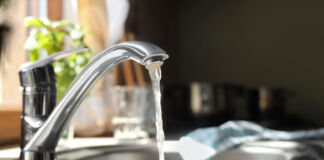 Ein Blick in eine Küche, direkt auf eine Spüle. Der Wasserhahn ist angeschaltet und es fließt klares Leitungswasser in das Waschbecken. Im Hintergrund kann man ein Geschirrhandtuch und einen Topf mit Küchenkräutern erkennen.