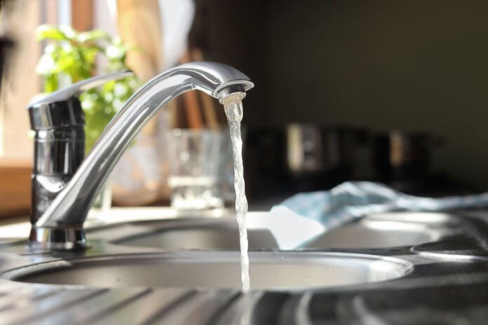 Ein Blick in eine Küche, direkt auf eine Spüle. Der Wasserhahn ist angeschaltet und es fließt klares Leitungswasser in das Waschbecken. Im Hintergrund kann man ein Geschirrhandtuch und einen Topf mit Küchenkräutern erkennen.