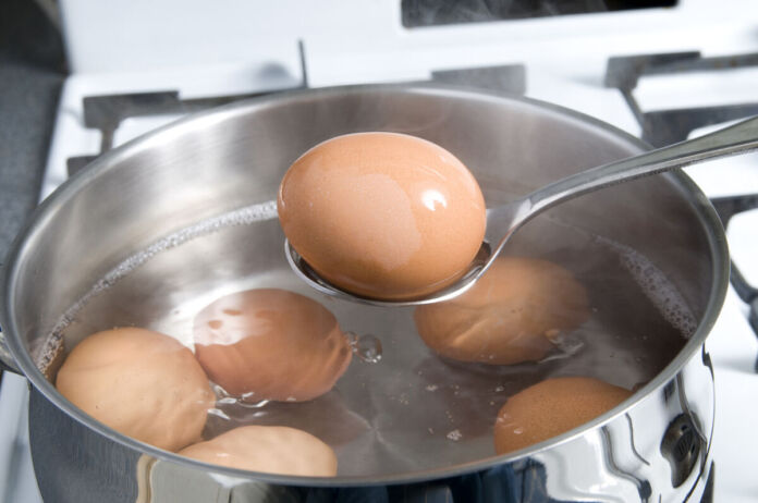 Sechs kochende Eier für das Frühstück in heißem Wasser in einem Kochtopf auf einem Küchenherd. Ein Ei wird auf einem Löffel platziert und herausgenommen.