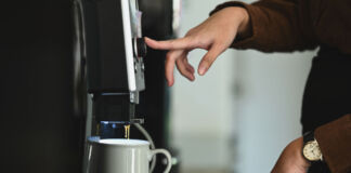 Eine Frau drückt gerade auf einen Knopf auf einer Kaffeemaschine, um einen Kaffee zu bekommen. An der entsprechenden Stelle steht eine Kaffeetasse. Die ersten Tropfen Kaffee laufen bereits aus der Maschine in die Tasse.