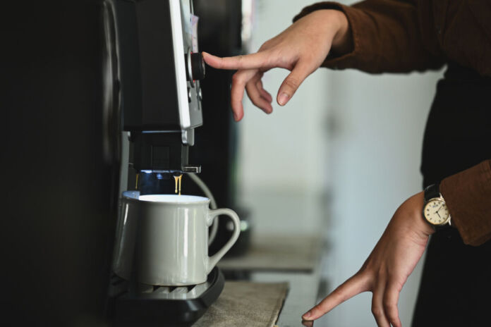 Eine Frau drückt gerade auf einen Knopf auf einer Kaffeemaschine, um einen Kaffee zu bekommen. An der entsprechenden Stelle steht eine Kaffeetasse. Die ersten Tropfen Kaffee laufen bereits aus der Maschine in die Tasse.