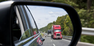 Das Bild von einem Rückspiegel eines Autos. In diesem erkennt man, dass der Pkw auf der Autobahn fährt. Hinter ihm befindet sich ein großer roter Lkw.