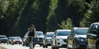 Eine glücklich aussehende Frau fährt Fahrrad auf der Straße. Neben ihr stehen viele Autos im Stau, während sie an ihnen vorbei pendelt und sie überholt. Dabei trägt sie einen Helm und lächelt.